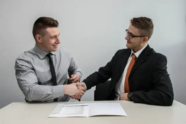 Dwóch mężczyzn w garniturach ściska sobie dłonie nad dokumentem, finalizując umowę przedwstępną sprzedaży nieruchomości.