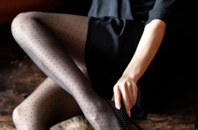 Kobieta w czarnej spódnicy i czarnych rajstopach w kropki zakłada buty na obcasie.