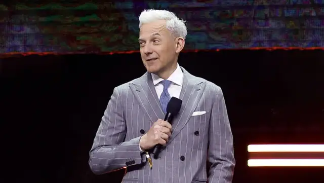 Mężczyzna w prążkowanym garniturze, prowadzący "Milionerzy", z mikrofonem w dłoni.