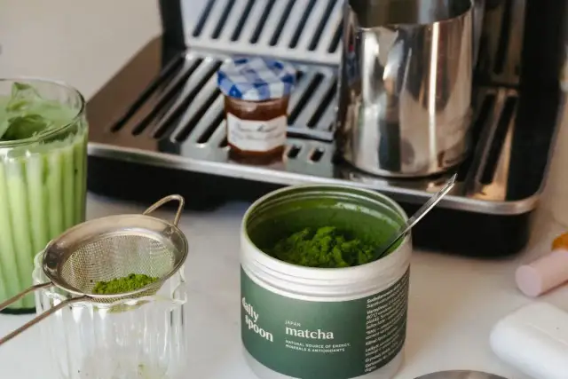 Puszka z zielonym proszkiem matcha, sitko z proszkiem, szklanka z napojem i ekspres do kawy.