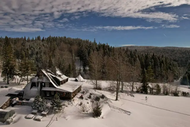 Zimowa sceneria wokół bacówki nad Wierchomlą. Śnieg pokrywa dachy, drzewa i okoliczne wzgórza.