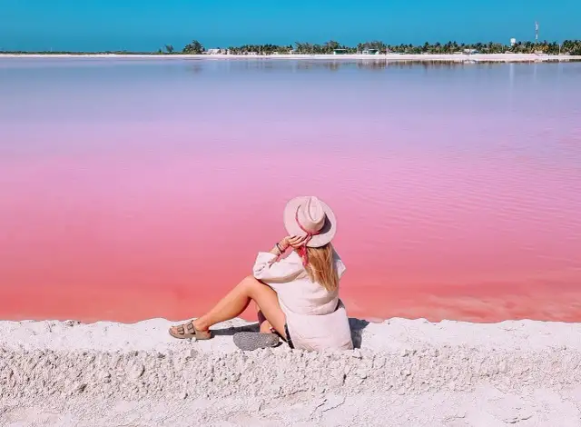 Kobieta w kapeluszu siedzi na piasku, podziwiając niezwykłą, różową plażę w Meksyku.