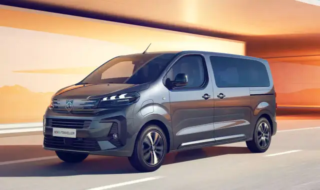 Szary bus Peugeot e-Traveller z przyciemnianymi szybami na tle pomarańczowej architektury.