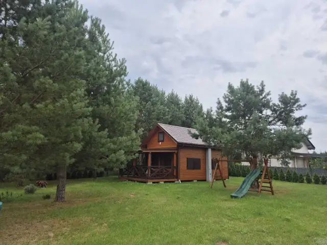Drewniany domek letniskowy otoczony drzewami, z werandą i zjeżdżalnią.