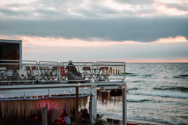 Widok na plażowy bar z widokiem na morze o zachodzie słońca, z ludźmi siedzącymi przy stolikach.