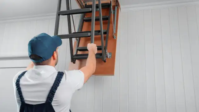 Mężczyzna w czapce i ogrodniczkach składa metalowe schody strychowe.