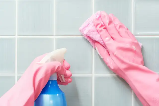 Ręce w różowych rękawiczkach czyszczą niebieskie kafelki sprayem i ściereczką.