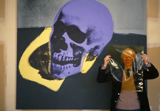 Andy Warhol trzyma przezroczystą folię przed fioletowo-żółtym obrazem czaszki.