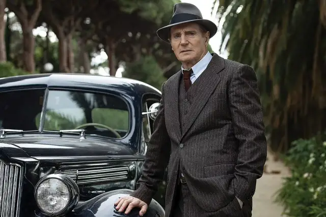 Philip Marlowe, detektyw z kryminałów Raymonda Chandlera, w eleganckim garniturze i kapeluszu, opiera się o luksusowy samochód z lat 40.