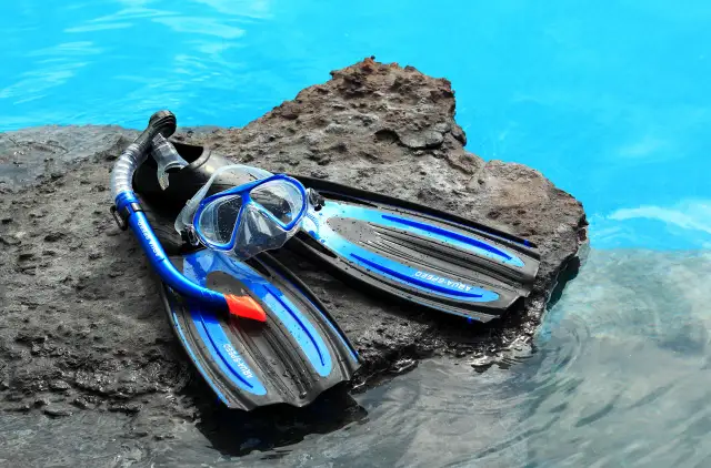 Idealne płetwy do nauki pływania: maska i rurka Aqua-Speed leżą na mokrej skale przy turkusowej wodzie.