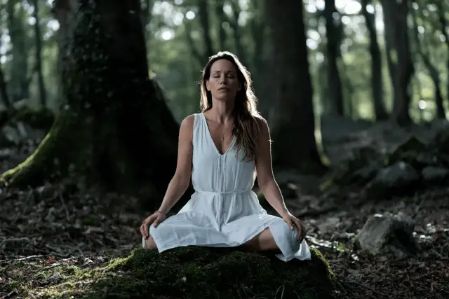 Kobieta w białej sukience medytuje w lesie na omszałym kamieniu.