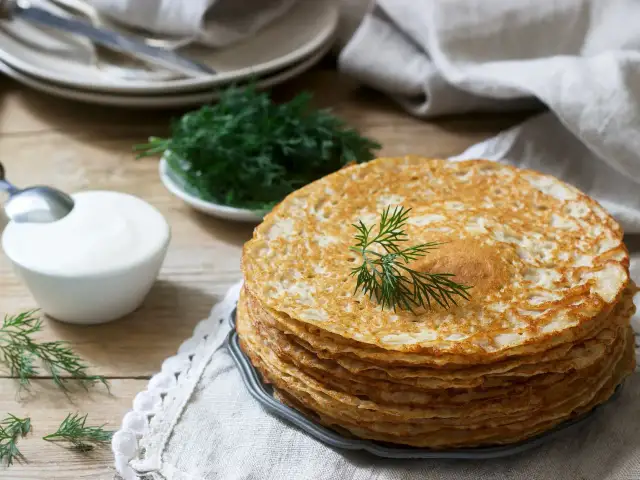Stos naleśników bezglutenowych, ozdobiony gałązką koperku, obok miseczki śmietany i świeżego koperku. Idealne na śniadanie.