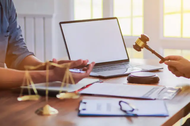 Dłoń trzymająca młotek sędziowski nad laptopem, obok waga sprawiedliwości.