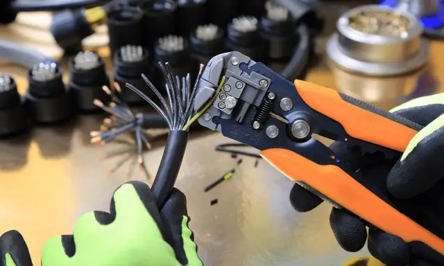 Ręka w rękawicy zdejmuje izolację z kabla za pomocą ściągacza.