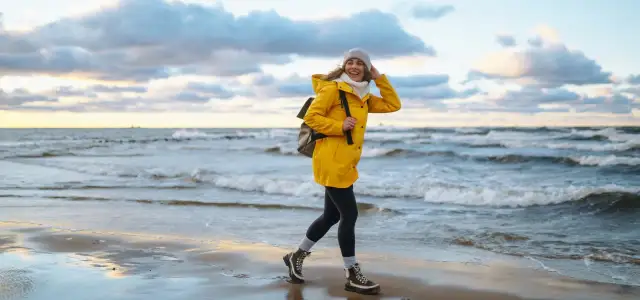 Uśmiechnięta kobieta w żółtej kurtce spaceruje po plaży. Wiatr rozwiewa jej włosy. To piękny jesienny dzień nad morzem.