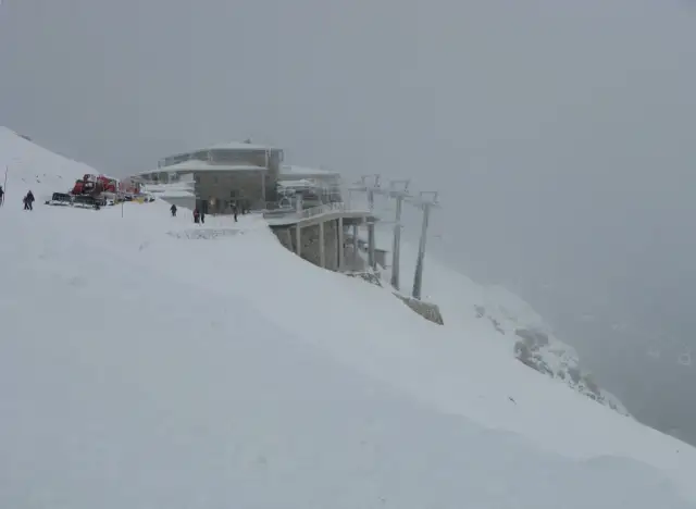 Mglisty dzień na Kasprowym Wierchu. Kolejka krzesełkowa wjeżdża na szczyt, a ratrak przygotowuje trasę do narciarstwa.