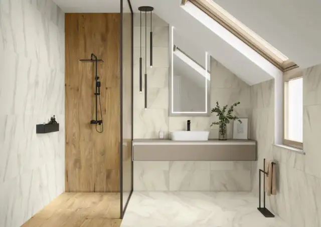 Nowoczesna, bardzo mała łazienka z prysznicem i pralką. Jasne płytki imitujące marmur, drewniana ściana w prysznicu, wisząca szafka z umywalką i podświetlane lustro.