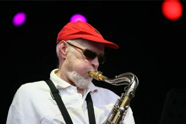 Starszy mężczyzna w czerwonej czapce i okularach gra na saksofonie.