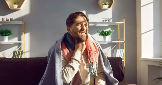 Mężczyzna z zarostem, okryty kocem i szalikiem, odczuwa ostry ból gardła przy przełykaniu, choć nie ma gorączki.