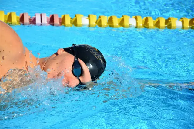 Pływak w czepku i okularach pokonuje wodę, pokazując, jak dobrze pływać kraulem. W tle widać żółte i różowe boje.