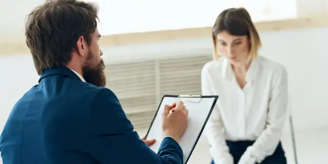 Mężczyzna z brodą w garniturze notuje coś na kartce, siedząc naprzeciwko kobiety.