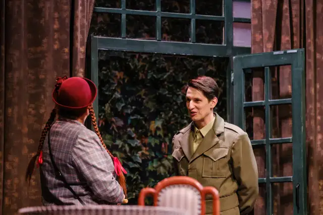 Na scenie młody mężczyzna w mundurze rozmawia z dziewczynką w czerwonym kapeluszu. W tle okno z zielonymi okiennicami. Widać, o co biega spektakl.