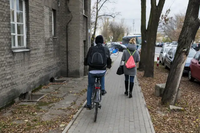 Osoba na rowerze i kobieta idąca chodnikiem obok budynku i drzew.