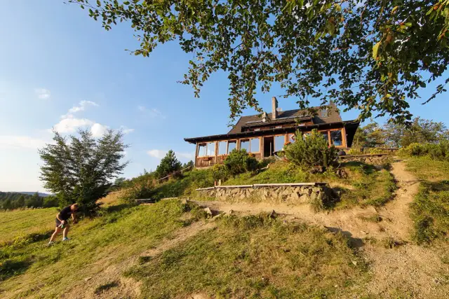 Bacówka PTTK nad Wierchomlą, drewniany dom z tarasem, otoczony zielenią, na zboczu wzgórza.