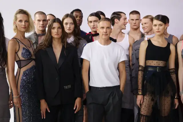 Grupa modeli i modelek pozuje obok słynnych projektantów mody. W centrum mężczyzna w białej koszulce, obok kobiety w czarnej marynarce.