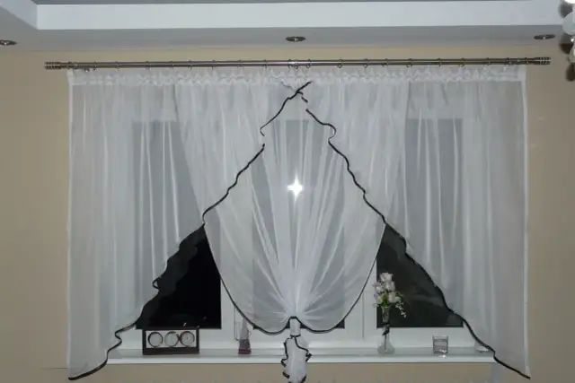Eleganckie białe firanki z brązową lamówką, idealnie pasujące na karnisz 2m. Na parapecie zegar i wazon z kwiatami.