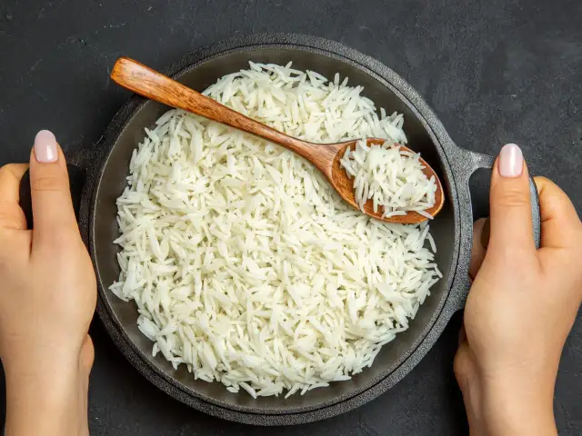 Sypki ryż jaśminowy w czarnej patelni z drewnianą łyżką, trzymanej przez dwie ręce.