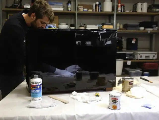 Mężczyzna w rękawiczkach czyści czarne, lakierowane meble. Na stole stoją puszki z preparatami i ściereczki.