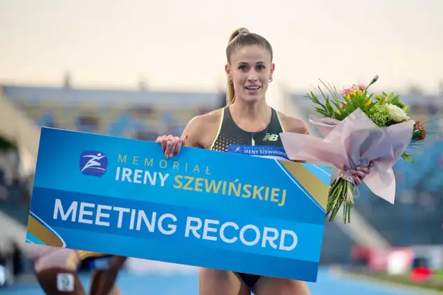 Uśmiechnięta lekkoatletka z bukietem kwiatów trzyma tablicę "MEETING RECORD" z logo memoriału Ireny Szewińskiej.
