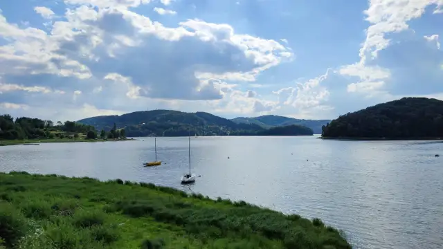 Widok na jezioro z żaglówkami i zalesionymi wzgórzami w tle, przy słonecznej pogodzie z chmurami.