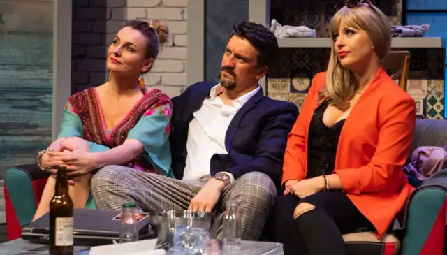 Trzy osoby siedzą na kanapie. Kobieta w kolorowej sukience, mężczyzna w marynarce i kobieta w pomarańczowej marynarce. Spektakl "Wiesz, że wiem".