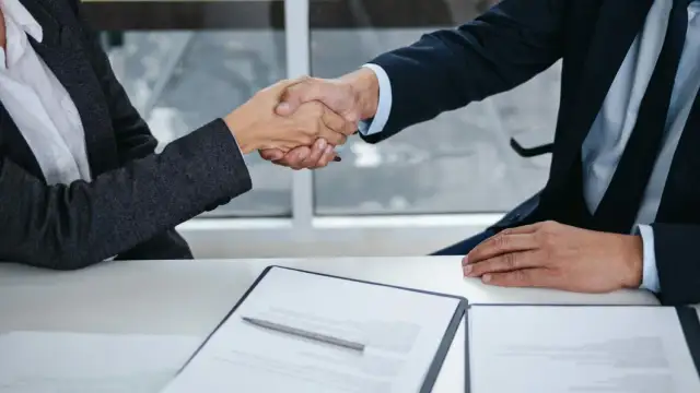 Dwie osoby w garniturach ściskają sobie dłonie nad stołem z dokumentami.