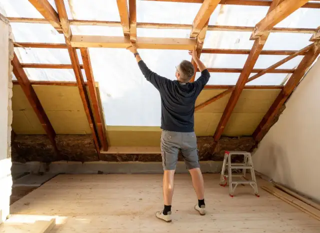 Mężczyzna montuje drewnianą konstrukcję poddasza, gdzie widać już wełnę mineralną. Zastanawiasz się, ile kosztuje metr ocieplenia poddasza?