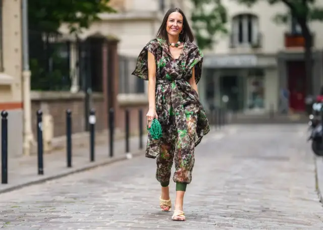 Kobieta w kwiecistym kombinezonie, zielonej torebce i plecionych sandałach. Idealna inspiracja, co założyć do kombinezonu.