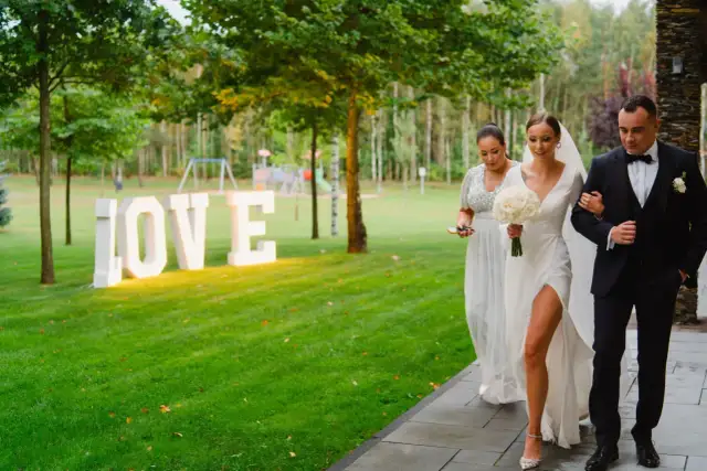 Panna młoda z bukietem idzie z panem młodym obok podświetlanych liter LOVE. Idealne tło do wierszyków na rocznice związku.