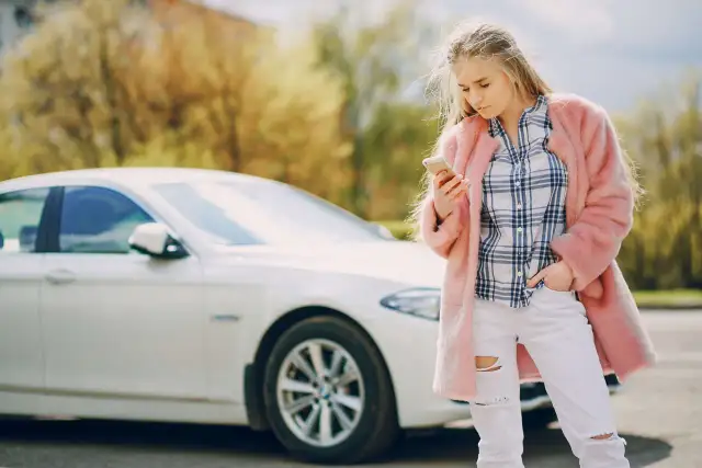 Młoda kobieta w różowym futrze i podartych dżinsach sprawdza telefon obok białego samochodu, być może po **szkodzie parkingowej**.