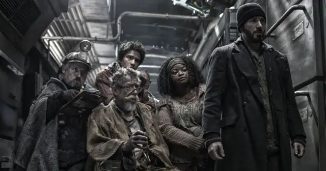 Grupa pasażerów Snowpiercer: Arka Przyszłości, w tym mężczyzna w czapce i płaszczu, siedzi w ciasnym wagonie.