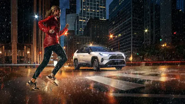Biegaczka w deszczowy wieczór w mieście, obok niej biała Toyota RAV4 Hybrid. Zastanawiasz się, ile pali Toyota RAV4 Hybrid?