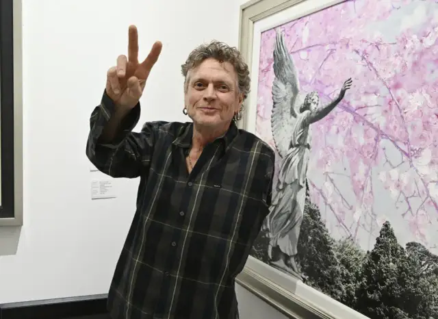 Rick Allen, perkusista Def Leppard, pozuje z uniesionym znakiem pokoju obok obrazu z aniołem i kwitnącymi drzewami.