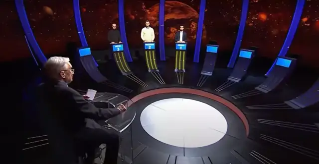 Trzech uczestników teleturnieju stoi przy swoich stanowiskach, a prowadzący siedzi przodem do nich.