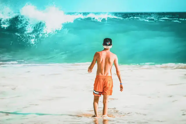 Mężczyzna w pomarańczowych spodenkach i czapce stoi w wodzie na tle dużej, turkusowej fali.