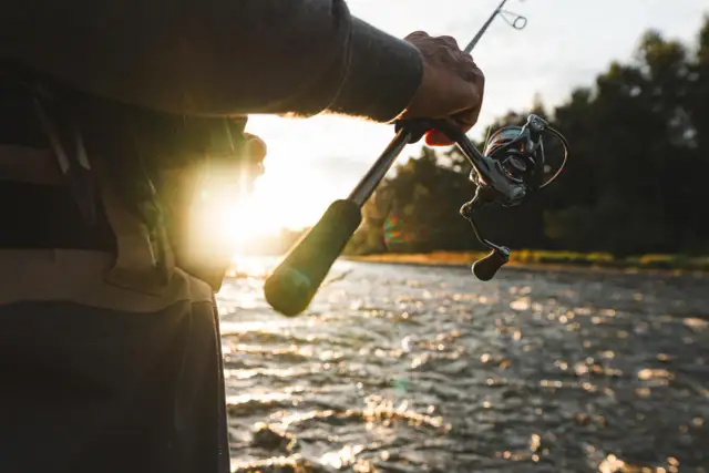 Wędkarz trzymający wędkę z kołowrotkiem nad wodą podczas zachodu słońca.