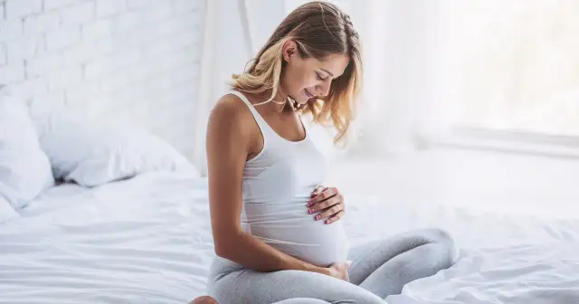 Uśmiechnięta kobieta w białej koszulce i szarych legginsach siedzi na łóżku, dotykając swojego twardego brzucha przed porodem.