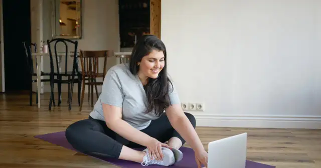 Uśmiechnięta kobieta w stroju sportowym siedzi na macie do ćwiczeń przed laptopem.