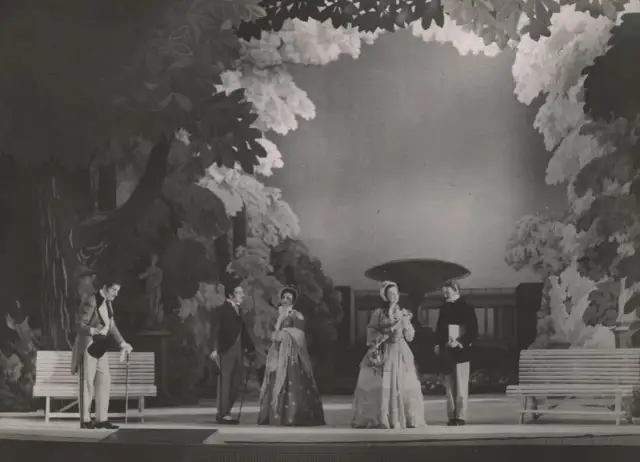 Scena z przedstawienia teatralnego, gdzie aktorzy w strojach z epoki odgrywają scenę w ogrodzie. Reżyser teatralny Szyfman zadbał o szczegóły scenografii.