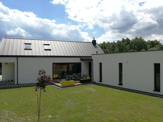 Nowoczesny, biały mały dom z płaskim dachem i dużymi oknami. Na tarasie stolik z krzesłami, w ogrodzie trawnik i młode drzewko.